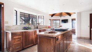 A spacious kitchen with wooden cabinets, a large island, a ceiling fan, and a TV mounted on the wall. - Cathedral by Avantstay Charming Secluded Estate w/ Sauna, Pool Table & Views of the Rolling Hills (Templeton)
