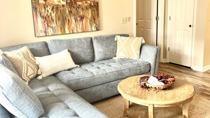 A living room with a grey sectional sofa, a round wooden coffee table, and a large painting of a cityscape. - LOCATION! | Walk to Stadium and Toomer’s | Cozy 4BR Condo in the Heart of Auburn (Auburn)