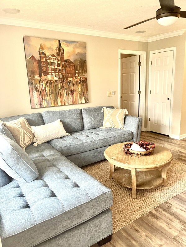 A living room with a grey sectional sofa, a round wooden coffee table, and a large painting of a cityscape.