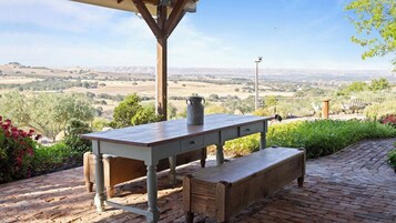 Un patio al aire libre con una mesa, un banco, un cubo de metal y vistas a un paisaje rural.