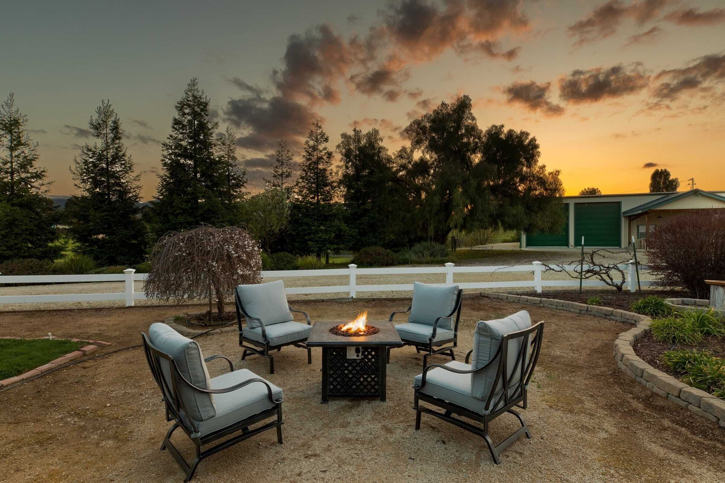 A patio with four chairs and a central fire pit, surrounded by a white fence and trees.