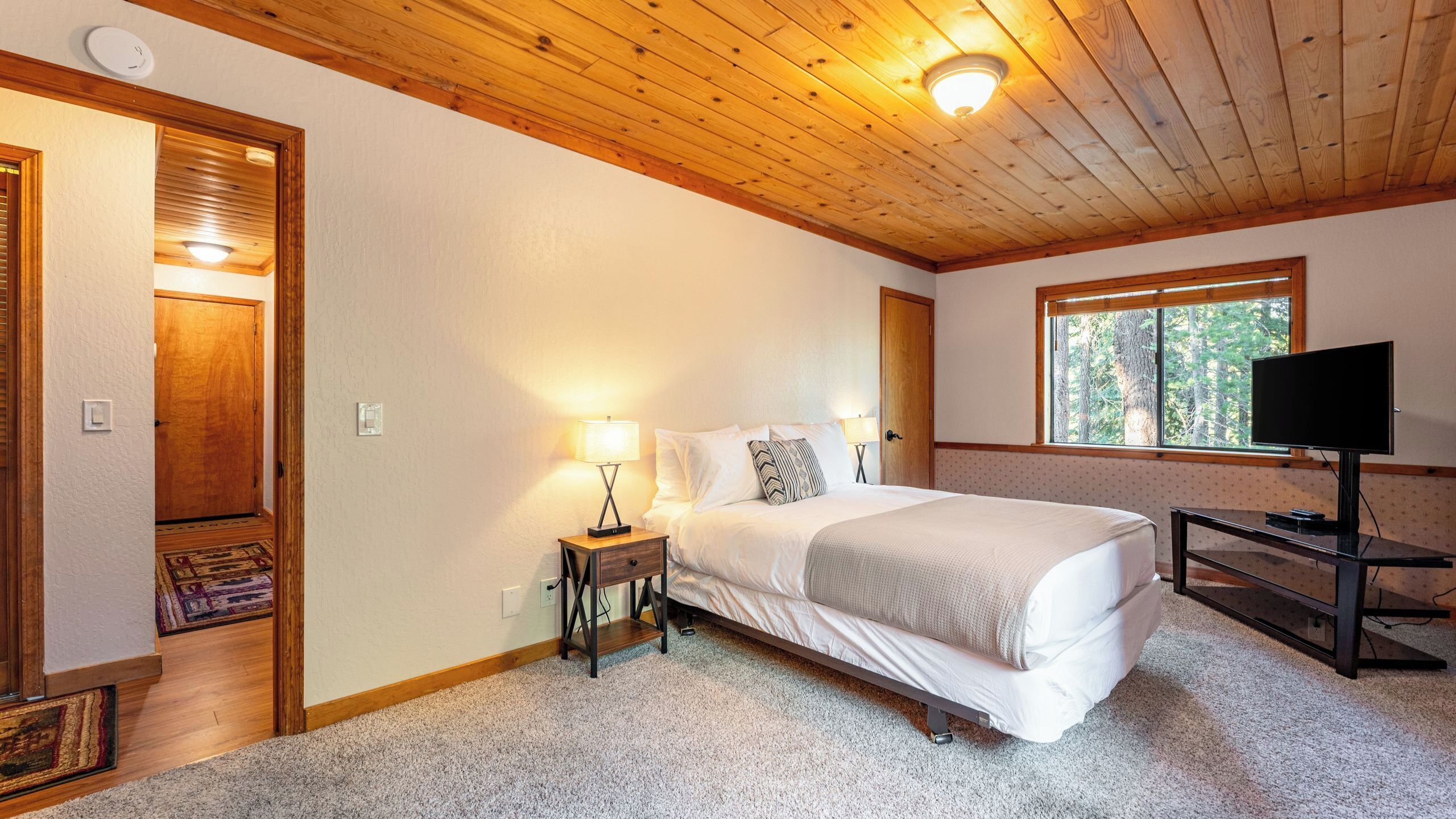 A bedroom with a bed, bedside lamps, a television, a window with a view of trees, and a wooden ceiling.