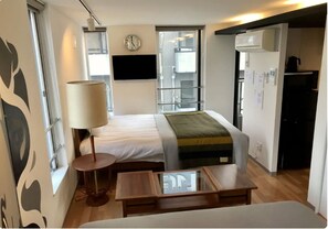 Une chambre d’hôtel de taille réduite, comprenant un lit, une table de chevet, une lampe, une fenêtre et une horloge fixée au mur.