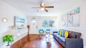 Un salon doté d’un parquet, d’un canapé gris agrémenté de coussins colorés, de fauteuils turquoise, d’une table basse blanche et d’un ventilateur de plafond.