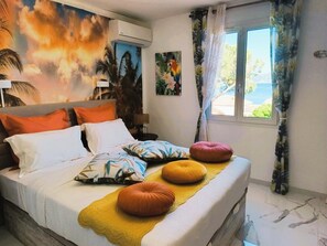 Un lit avec plusieurs oreillers, une peinture murale, une fenêtre avec des rideaux et une lampe.