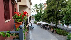 A street with outdoor seating, a building with red shutters, and a row of trees.