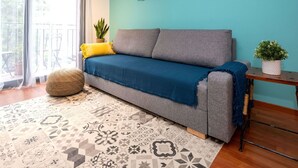 A living room with a grey sofa, a blue throw blanket, a patterned rug, a small wooden side table with a plant, and a window with sheer curtains.