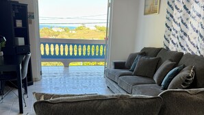 A living room with a grey sofa, a small table, and a chair. There is a door leading to a balcony with a view of the countryside. - Ocean View Home with private pool and parking. Sleeps 13 (Rincon)