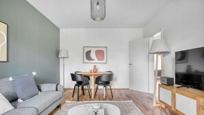 Un salon moderne avec un canapé gris, une table basse ronde sur laquelle repose un livre, un coin repas avec une table et des chaises, un téléviseur posé sur un meuble en bois et un luminaire suspendu.