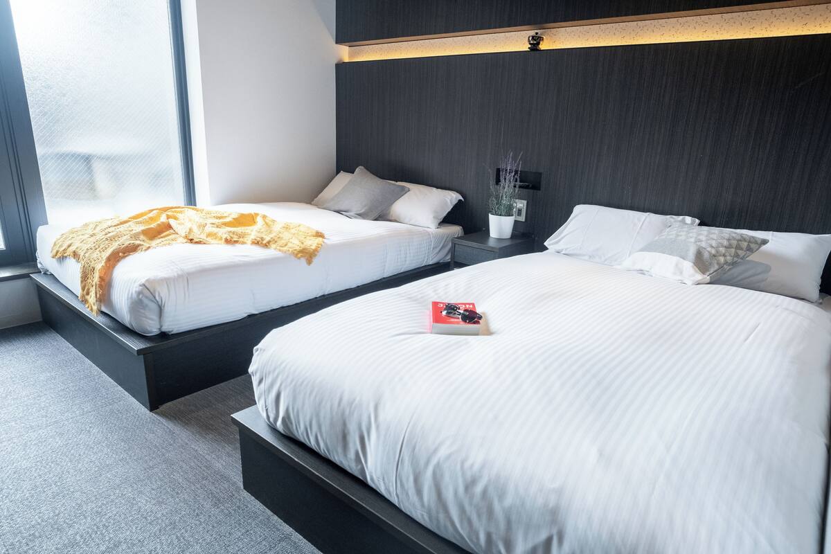 A modern hotel room with two beds, a small bedside table with a potted plant, and a window providing natural light.