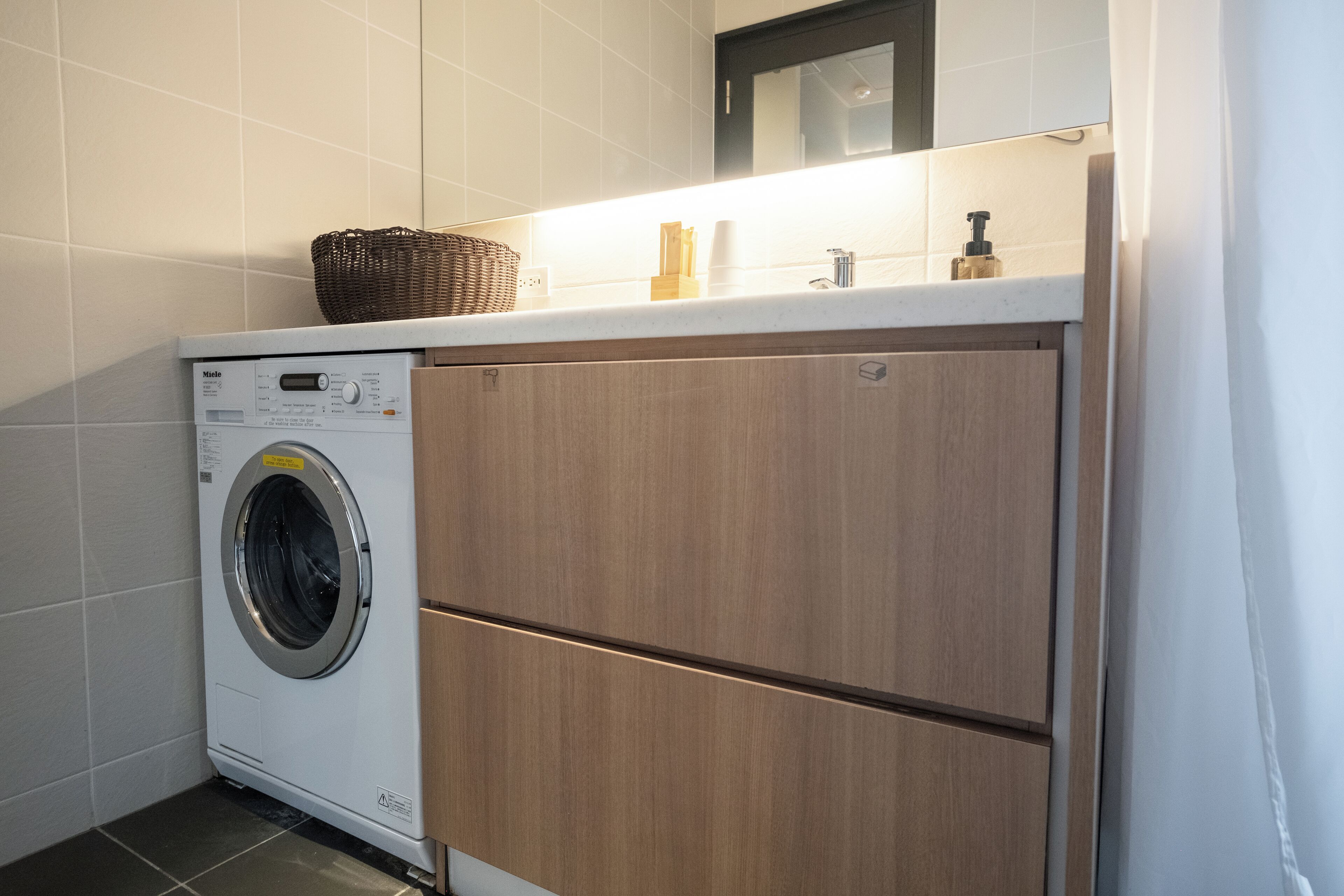 Un espace buanderie compact comprenant un lave-linge à chargement frontal, des placards en bois et un plan de travail avec un panier et des articles de toilette.