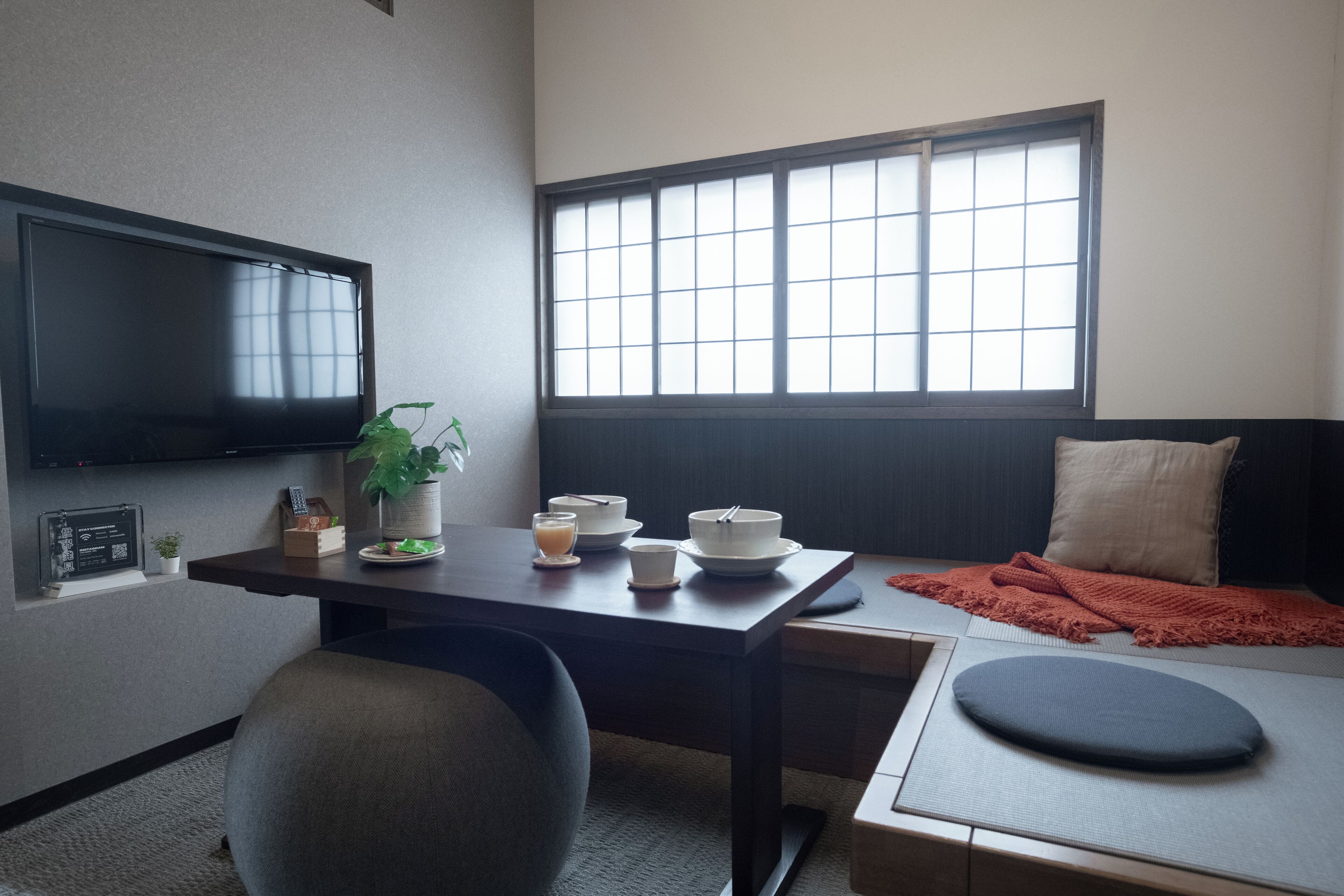 Una habitación compacta con un televisor montado en la pared, una mesa de comedor con cuencos y una planta, una cama con una manta y una ventana con persianas.