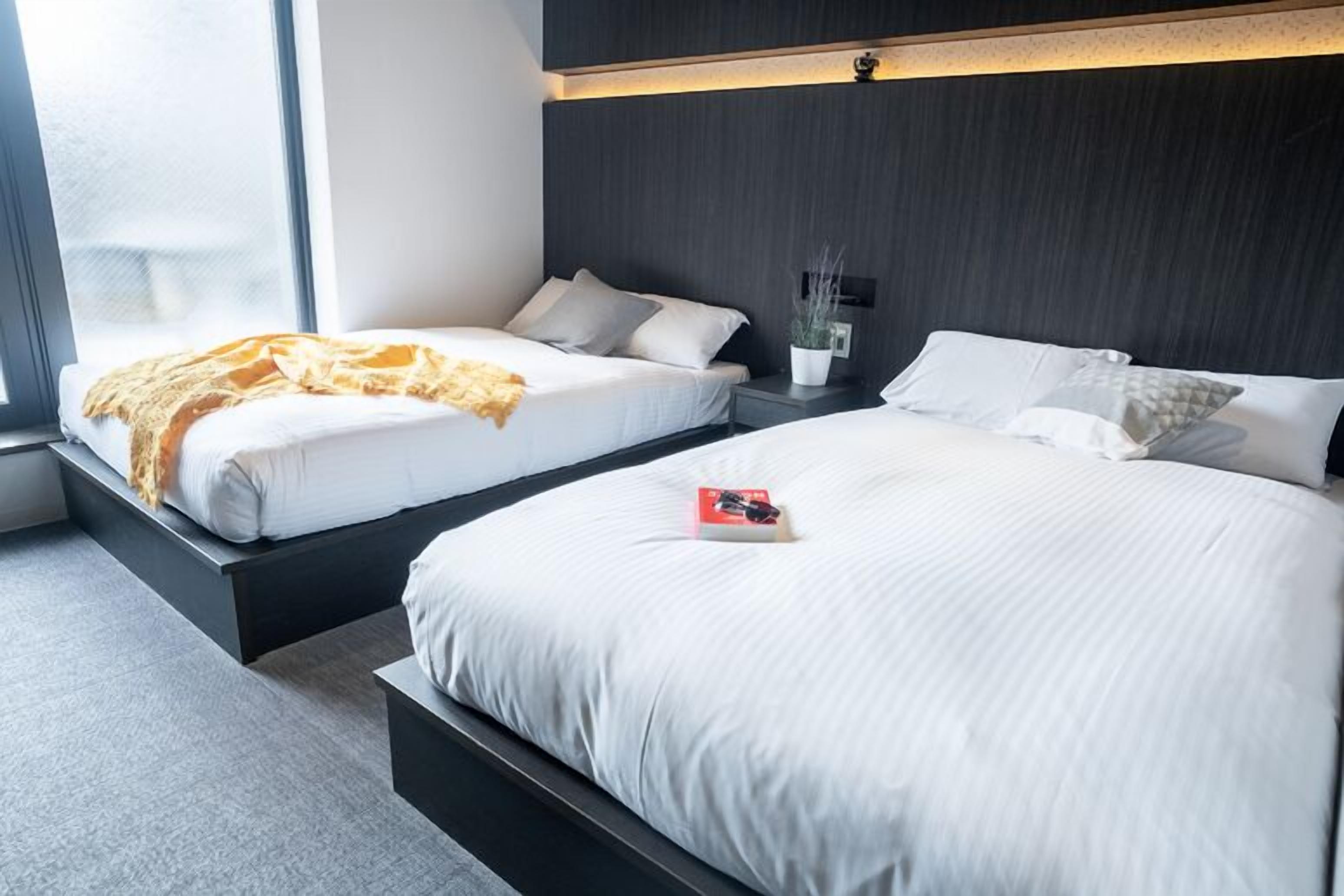A modern hotel room with two beds, a bedside table with a lamp, a window, and a view of the outside.