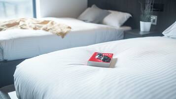 A hotel room with two beds, a bedside table with a plant, and a window providing natural light.