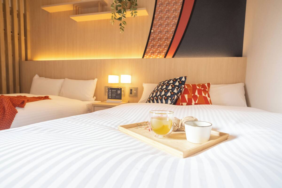 A hotel room with a large bed, a bedside table with a lamp, a tray with a cup and a glass of tea, and a wall with a geometric pattern.
