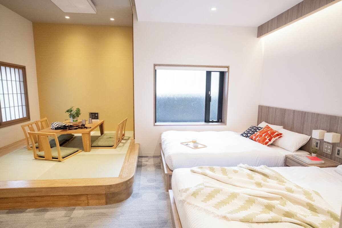 A hotel room with two beds, a dining area with a table and chairs, a window with a view, and a bedside table with a lamp.