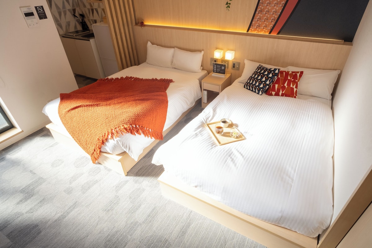 A hotel room with two beds, a small bedside table with a lamp, and a window.