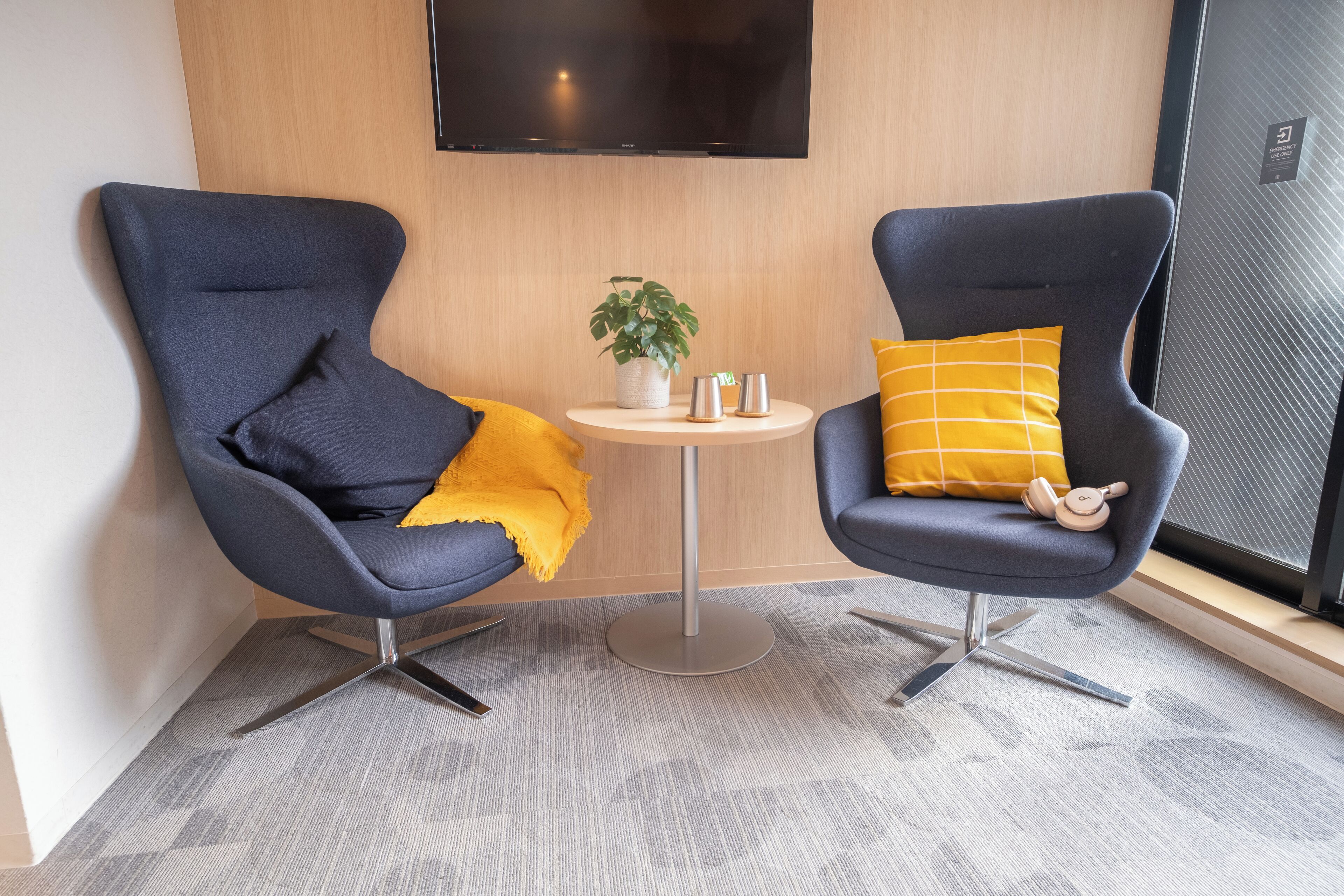 Two modern chairs with metal bases, a small round table with a plant and two cups, and a large flat-screen TV mounted on the wall.
