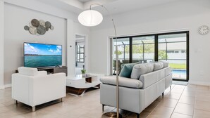 Un salon moderne doté d’un grand téléviseur à écran plat, de canapés blancs, d’un lampadaire et offrant une vue sur une piscine grâce à de grandes portes vitrées.
