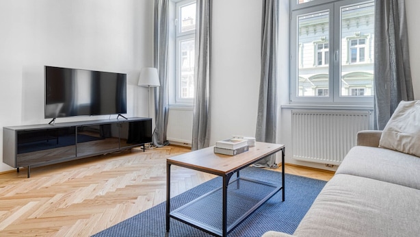 Ett modernt vardagsrum med en platt-TV, ett soffbord i trä, en soffa och utsikt över byggnader genom stora fönster.
