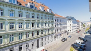 En byggnad med flera våningar, med en rad fönster i bottenvåningen. Till höger står en vit byggnad med ett blått tak, och längs gatan står parkerade bilar.
