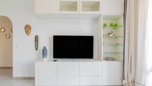 Una sala moderna con un televisor de pantalla plana sobre un mueble blanco, una estantería de vidrio con objetos decorativos y una pared lisa con un diseño geométrico.