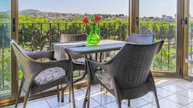 Un balcón con mesa y sillas, un jarrón verde con flores, y vistas a un viñedo y edificios al fondo.