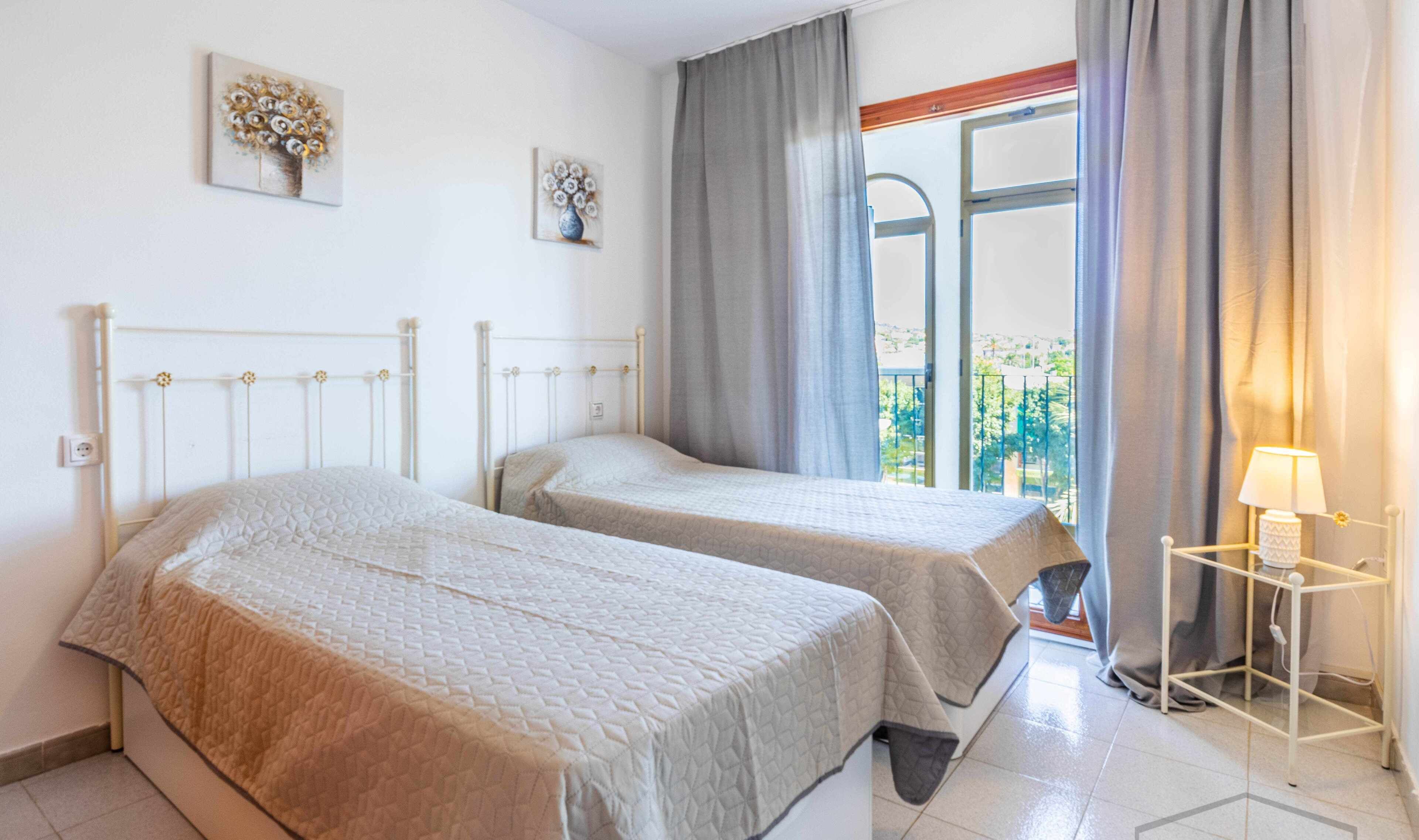 A hotel room with two beds, a window with curtains, a bedside table with a lamp, and artwork on the wall.