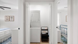 Un pasillo estrecho con una puerta blanca que da a un lavadero con lavadora. A la izquierda, una cama con colcha azul y blanca, y a la derecha, un pasamanos azul.