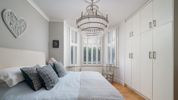 A bedroom with a large bed, a chandelier, white cabinets, and a window with shutters. - Gorgeous flat with garden in Battersea (London)