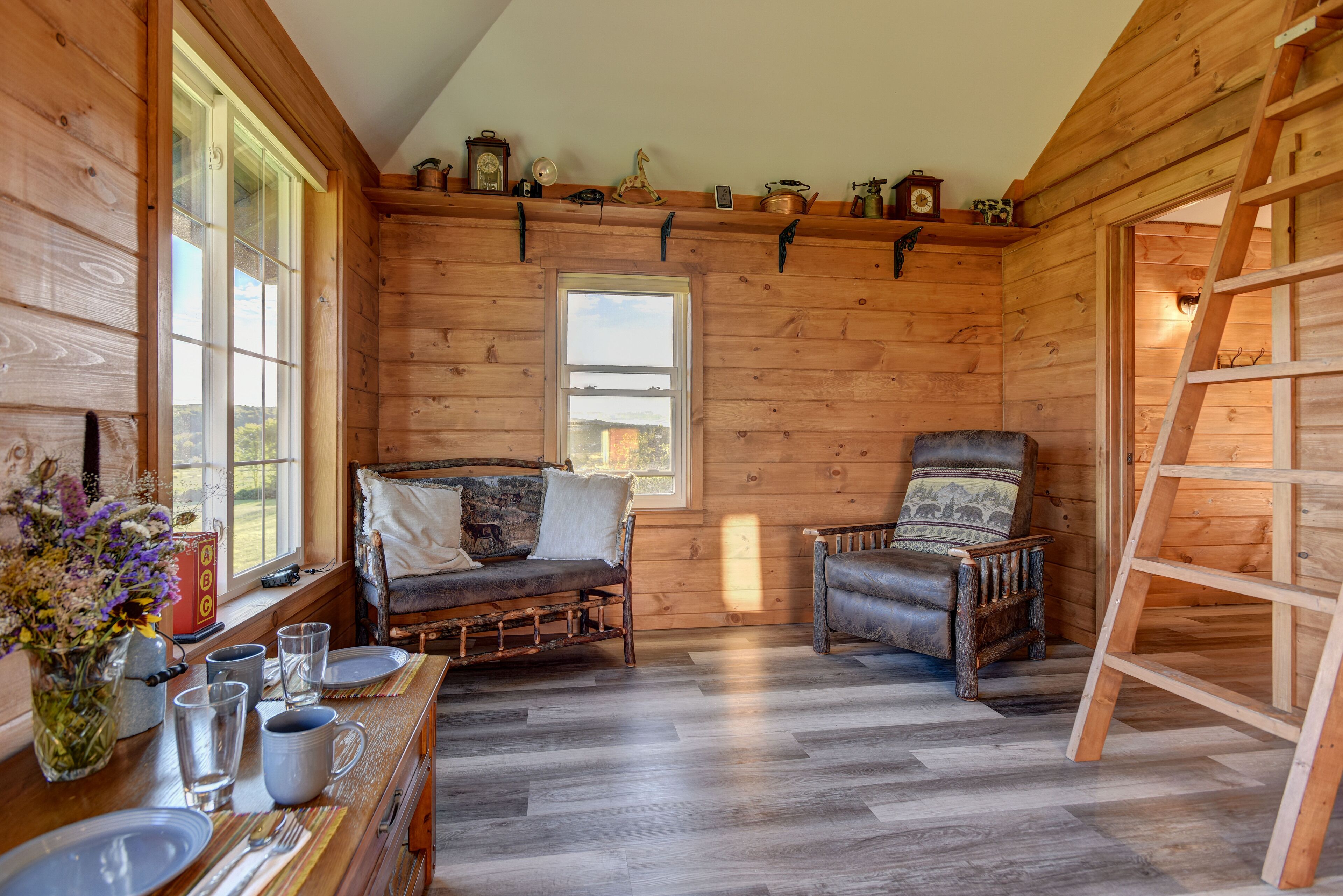 Interior acogedor de una cabaña de troncos con una mesa de comedor de madera dispuesta para dos personas, un banco acolchado, un sillón de cuero y una escalera apoyada en la pared.