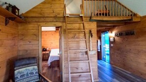 A wooden cabin interior with a loft bedroom, a ladder to the upper bunk, a chair with a patterned cushion, and a visible door leading to another room. - The Caboose | Mtn Views | Farm Animals + Sauna (Canajoharie)