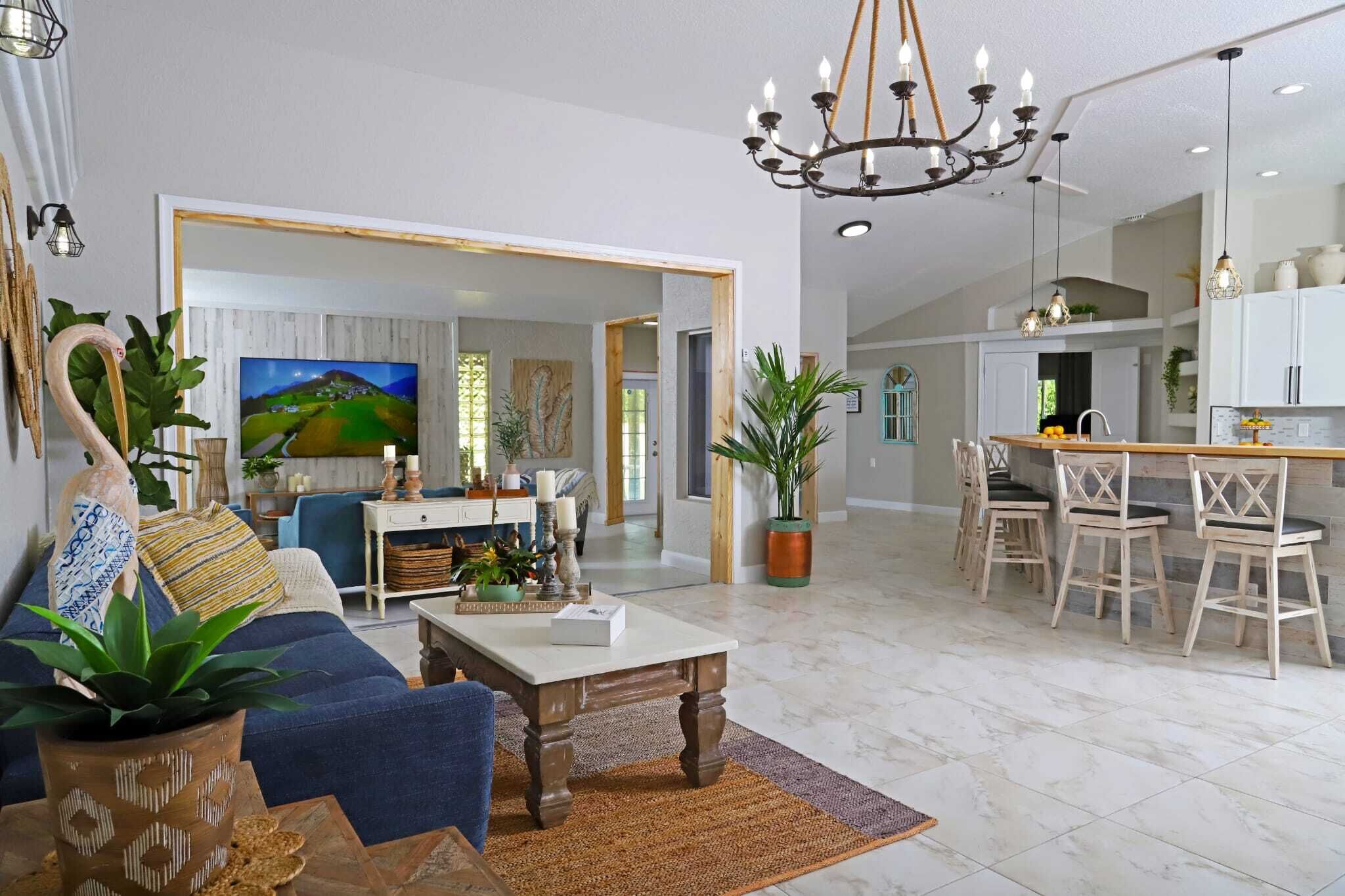 A spacious living room with a sofa, coffee table, and potted plants. A dining area with chairs and a kitchen in the background.
