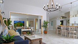 A spacious living room with a sofa, coffee table, and potted plants. A dining area with chairs and a kitchen in the background.