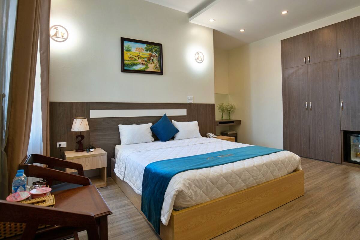 A hotel room with a large bed, a desk, a chair, a nightstand, a lamp, a painting, a wardrobe, and a minibar.