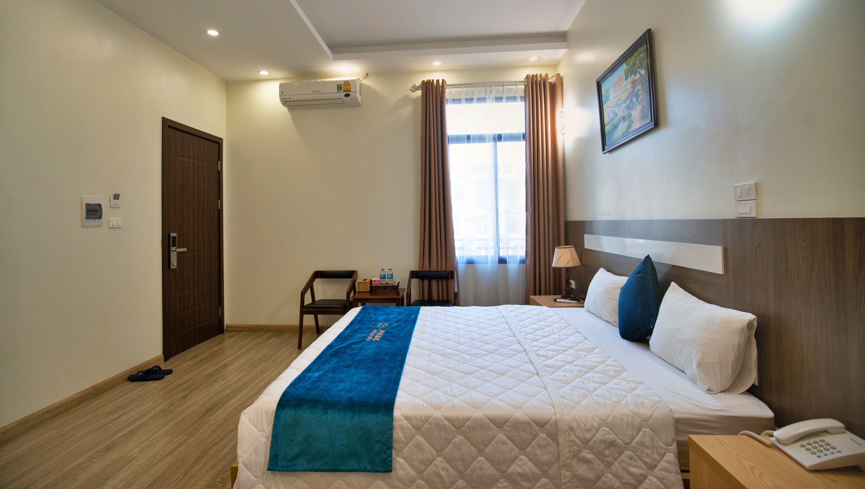 A hotel room with a bed, a desk with a phone, a chair, a window with curtains, an air conditioning unit, and a painting on the wall.