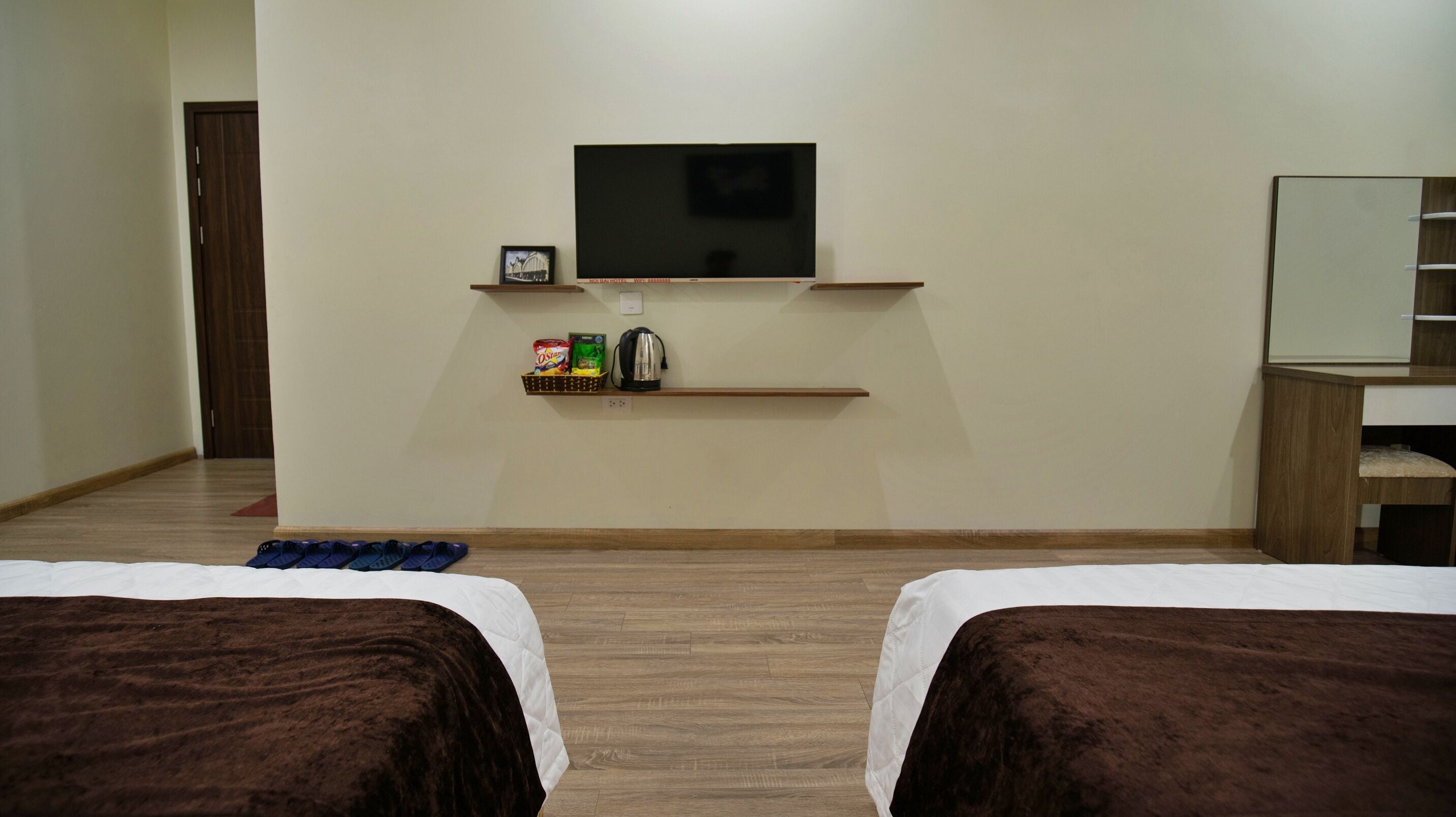A hotel room with two beds, a TV mounted on the wall, a shelf with toiletries, and a wooden floor.