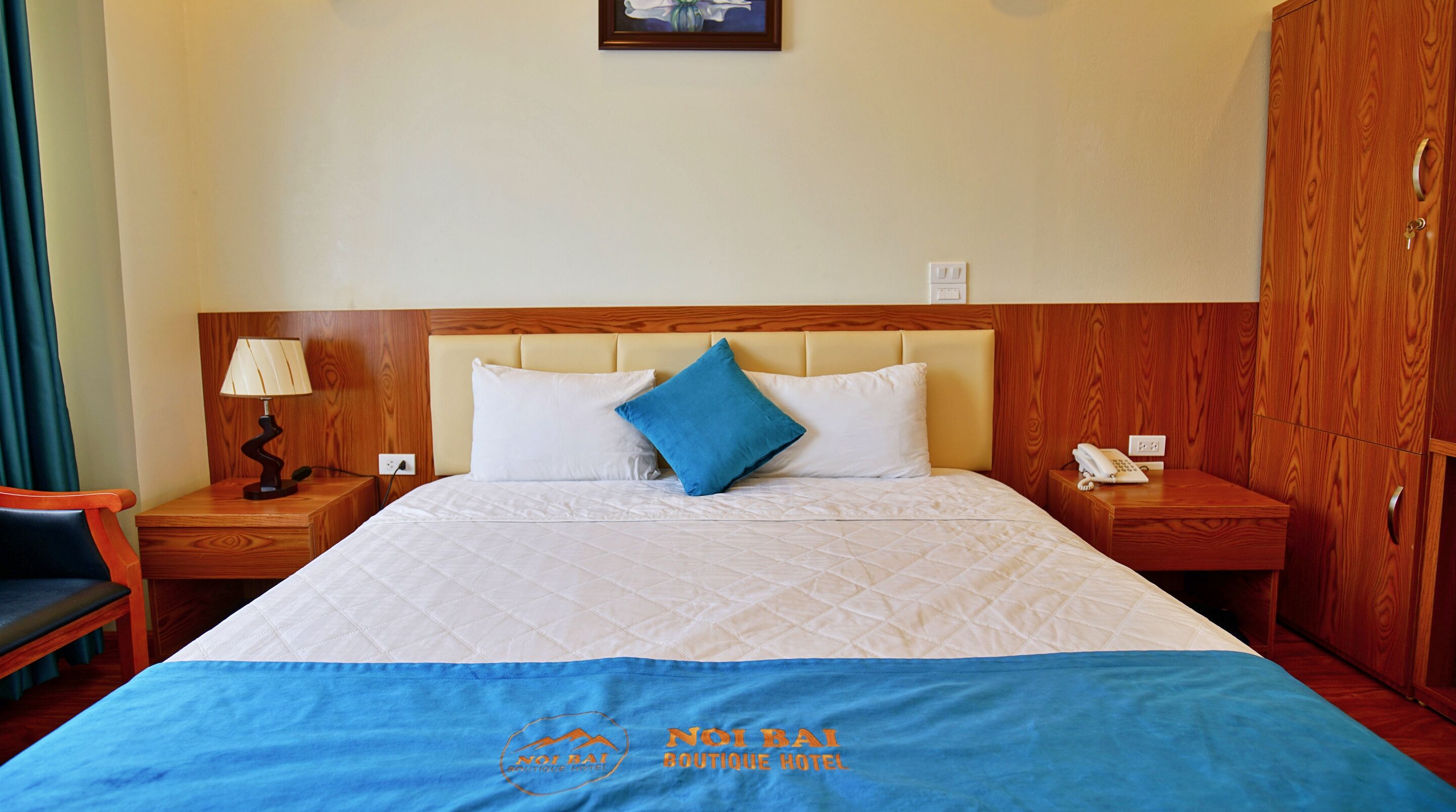 A hotel room with a large bed, a wooden headboard, a nightstand with a lamp, a telephone, and a wardrobe.