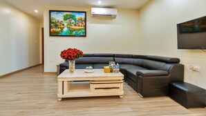 A living room with a black leather sofa, a wooden coffee table with a vase of red flowers, a flat-screen TV mounted on the wall, and an air conditioning unit. - S79 Noi Bai Airport Hotel (Hanoi)