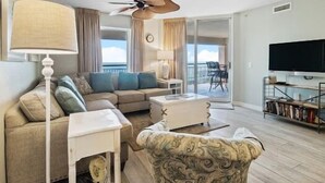 Een woonkamer met een bank, een gestreept fauteuil, een salontafel, een lamp, een plafondventilator, een televisie en een balkon met uitzicht.