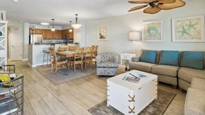Un salon avec un canapé, une table basse, un coin repas avec des chaises, une cuisine à l’arrière-plan et un ventilateur de plafond.