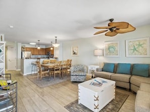 A living room with a sofa, a coffee table, a dining area with chairs, a kitchen in the background, and a ceiling fan.