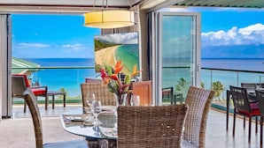 Uma varanda com móveis de vime, uma mesa de vidro e vista para o oceano.