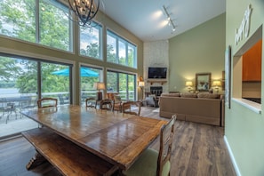 Un vaste espace de vie comprenant une grande table à manger en bois, des sièges confortables, une cheminée et de grandes fenêtres offrant une vue sur l’extérieur.