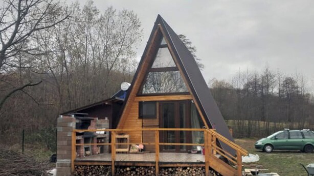 A triangular wooden cabin with a balcony, a staircase, and a car parked nearby.