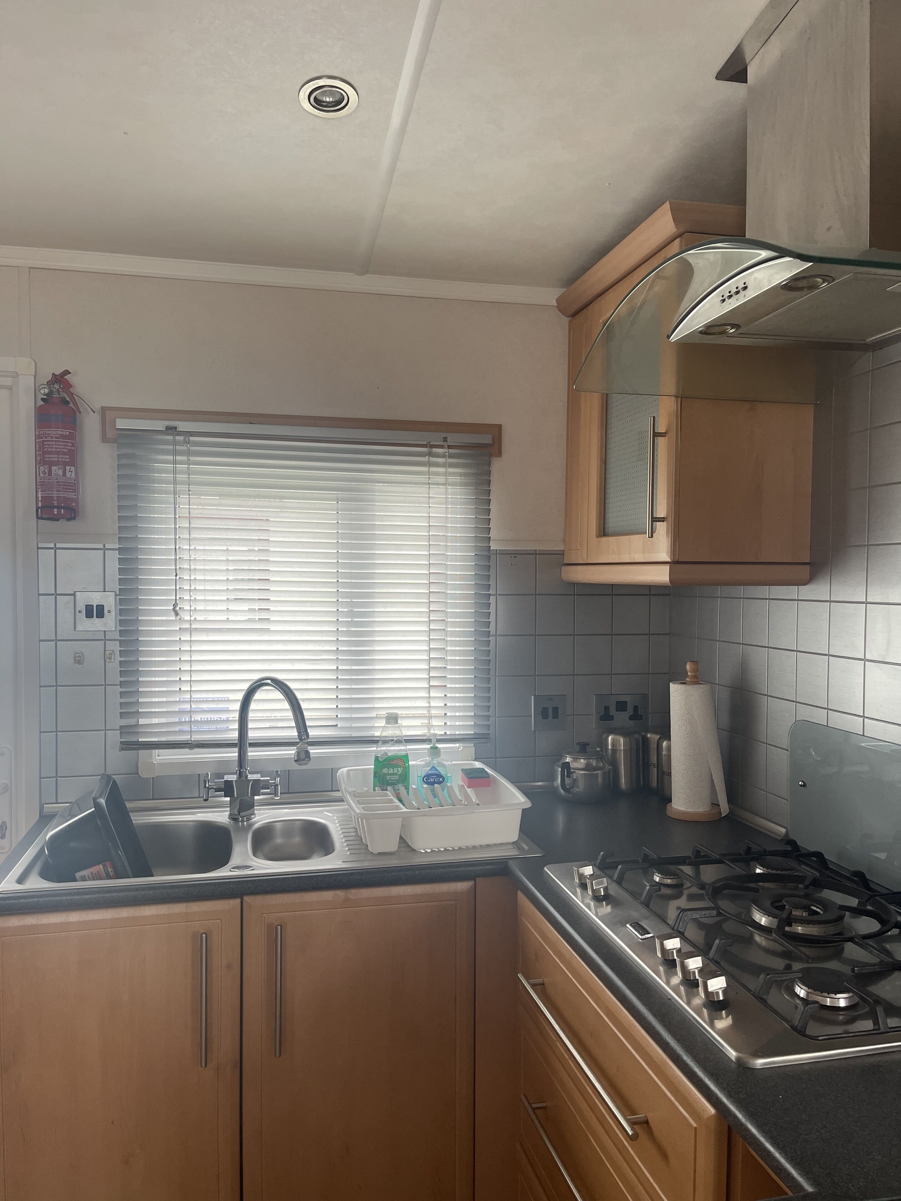 A kitchen with a gas stove, a sink, wooden cabinets, a window with blinds, and a fire extinguisher.