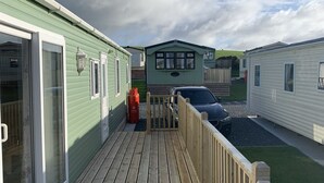 A wooden deck leading to a row of mobile homes with large windows and a grassy yard. - Immaculate 2-Bed Lodge in Monreith (Newton Stewart)