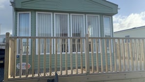 A single-story mobile home with a green exterior and white trim, featuring a balcony with a wooden railing. - Immaculate 2-Bed Lodge in Monreith (Newton Stewart)