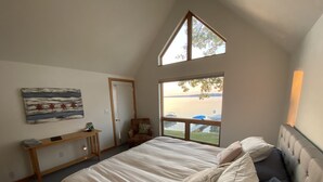 Une chambre avec un grand lit, un bureau, une chaise, une fenêtre donnant sur un plan d’eau et un tableau accroché au mur.