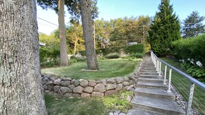 Un escalier en pierre menant à une maison, entourée d’un mur de pierre et d’une zone herbeuse avec des arbres.